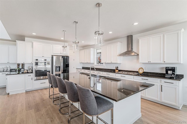 kitchen featuring a center island with sink, appliances with stainless steel finishes, wall chimney exhaust hood, and light wood-style floors