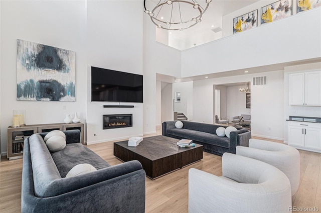 living area with visible vents, an inviting chandelier, a towering ceiling, stairs, and light wood-style flooring