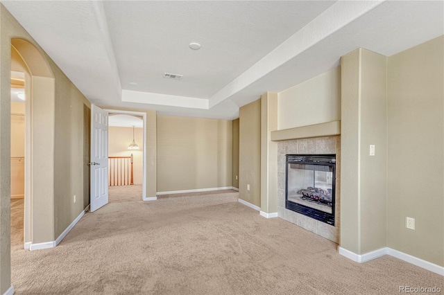 unfurnished living room with arched walkways, light colored carpet, visible vents, a tray ceiling, and a tiled fireplace