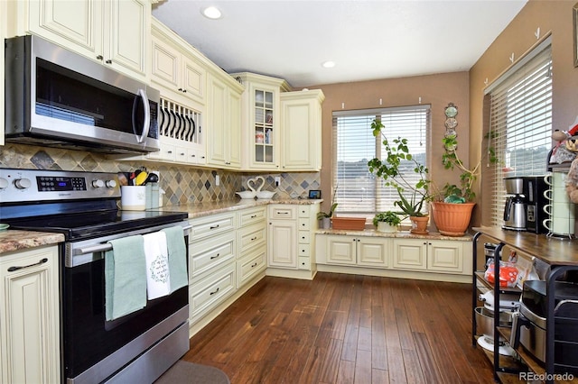 kitchen with light stone counters, cream cabinetry, appliances with stainless steel finishes, dark wood-style floors, and glass insert cabinets