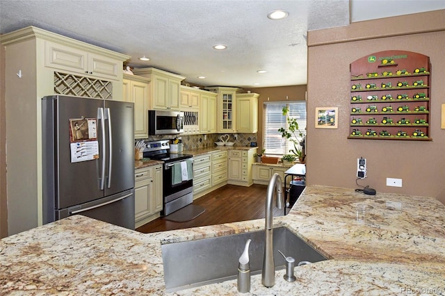 kitchen with cream cabinetry, appliances with stainless steel finishes, glass insert cabinets, and light stone countertops