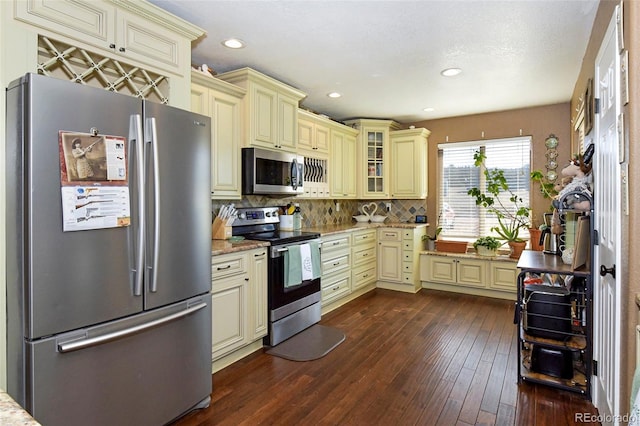 kitchen featuring stainless steel appliances, cream cabinets, glass insert cabinets, and light stone countertops