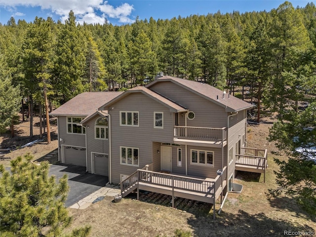 back of property with an attached garage, a view of trees, asphalt driveway, and a balcony