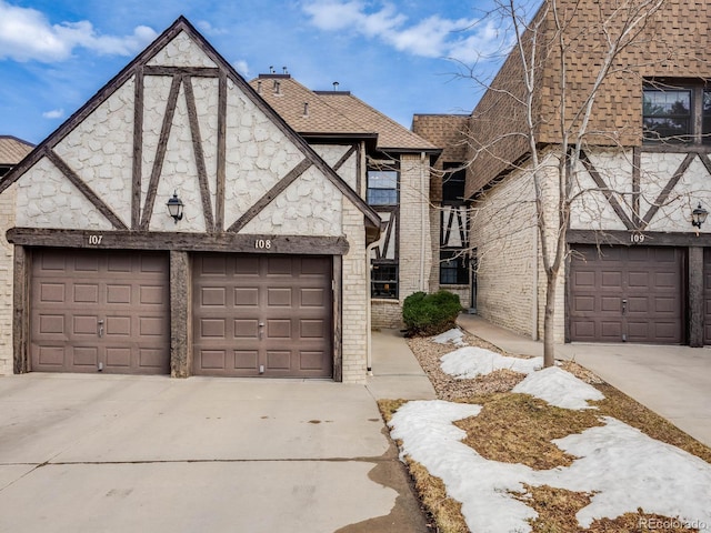 tudor house with roof with shingles, brick siding, a garage, stone siding, and driveway