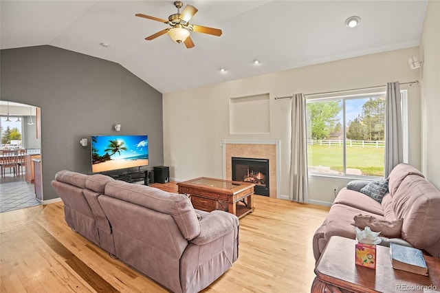 living room with ceiling fan, light wood-type flooring, a tile fireplace, and vaulted ceiling