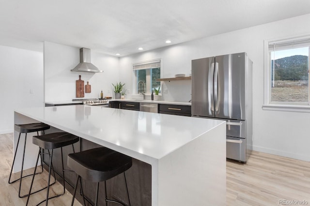 kitchen with stainless steel appliances, a center island, wall chimney range hood, and light wood-type flooring