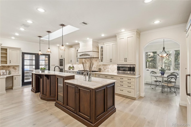 kitchen featuring pendant lighting, glass insert cabinets, appliances with stainless steel finishes, an island with sink, and premium range hood