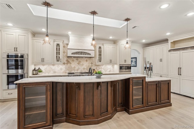 kitchen with premium range hood, a spacious island, hanging light fixtures, appliances with stainless steel finishes, and glass insert cabinets