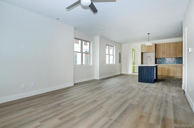 kitchen with backsplash, light hardwood / wood-style flooring, ceiling fan, an island with sink, and decorative light fixtures