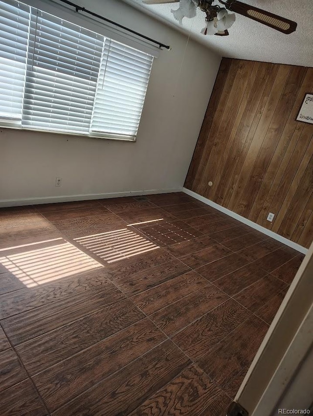 unfurnished room with a textured ceiling, dark wood-style floors, wood walls, baseboards, and ceiling fan