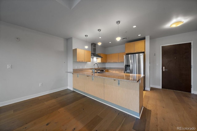 kitchen with kitchen peninsula, stainless steel refrigerator with ice dispenser, hanging light fixtures, light brown cabinets, and wall chimney exhaust hood