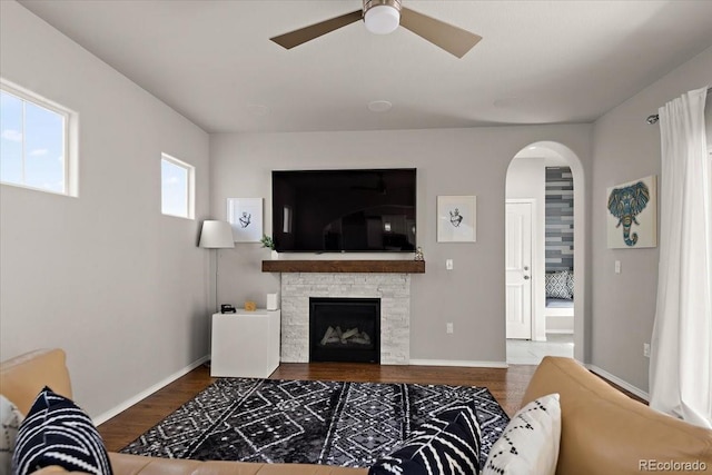 living room with arched walkways, a ceiling fan, a stone fireplace, wood finished floors, and baseboards