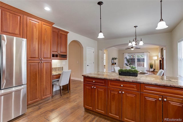 kitchen with light stone counters, wood finished floors, freestanding refrigerator, brown cabinets, and decorative light fixtures