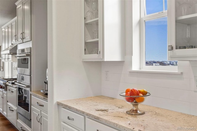kitchen with tasteful backsplash, glass insert cabinets, stainless steel appliances, white cabinets, and light stone countertops