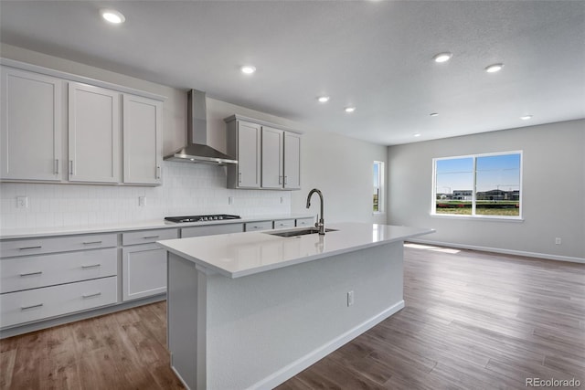 kitchen with wood finished floors, a center island with sink, a sink, wall chimney exhaust hood, and tasteful backsplash