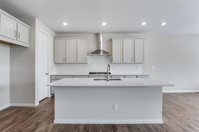 kitchen with baseboards, a kitchen island with sink, a sink, wall chimney exhaust hood, and tasteful backsplash