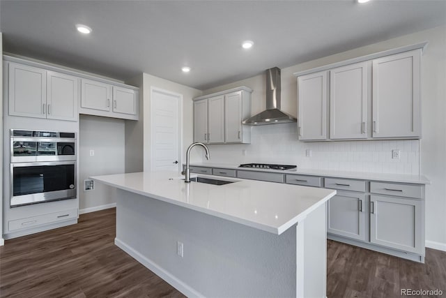 kitchen with a sink, backsplash, appliances with stainless steel finishes, wall chimney exhaust hood, and dark wood-style flooring