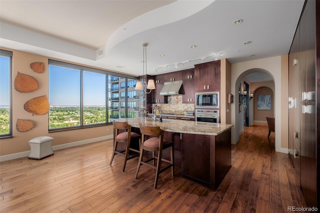 kitchen with built in microwave, oven, hardwood / wood-style floors, light stone countertops, and a kitchen island with sink