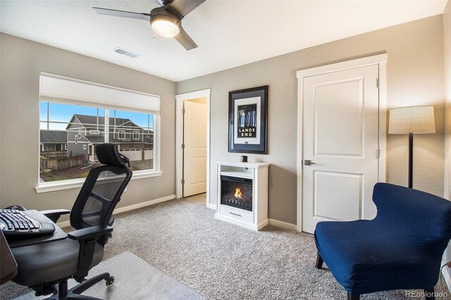 office space with a warm lit fireplace, ceiling fan, carpet, baseboards, and visible vents