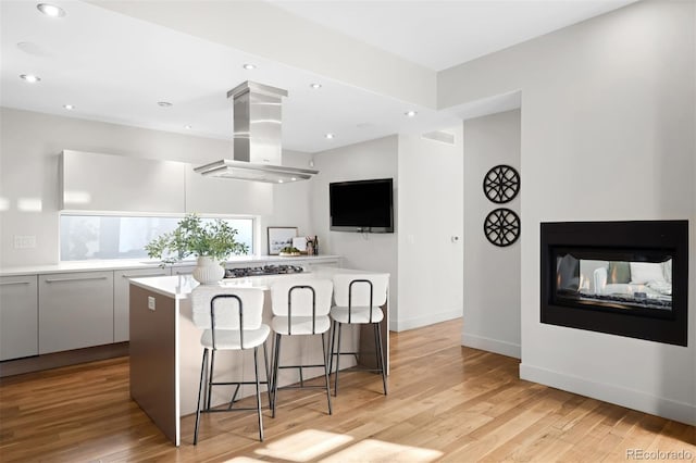 kitchen with island exhaust hood, a breakfast bar area, light wood finished floors, light countertops, and a multi sided fireplace