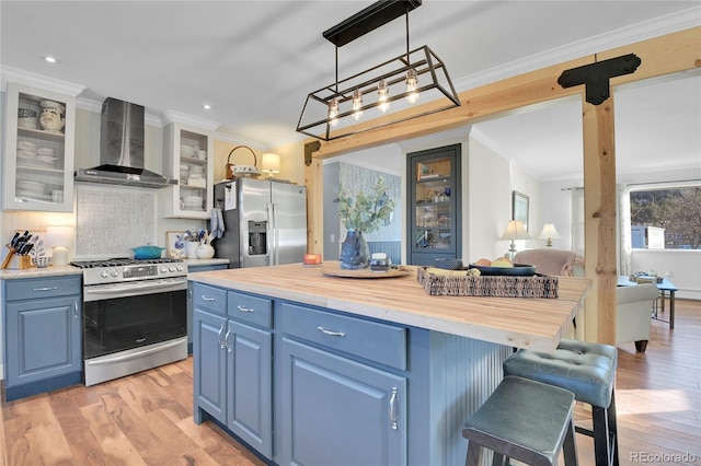 kitchen with wooden counters, appliances with stainless steel finishes, blue cabinets, pendant lighting, and wall chimney exhaust hood