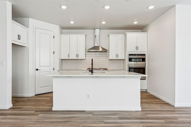 kitchen featuring stainless steel double oven, white cabinets, an island with sink, and wall chimney exhaust hood