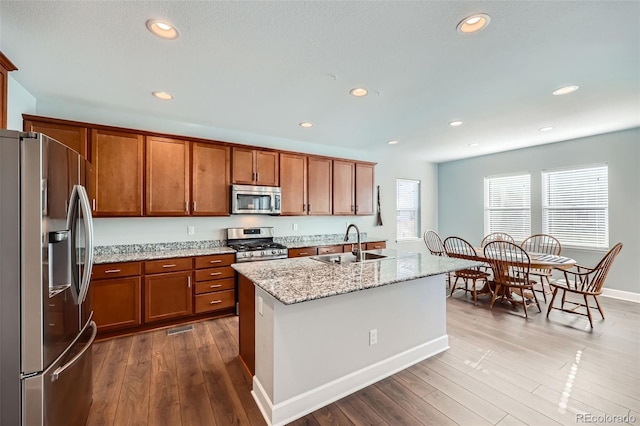 kitchen featuring sink, hardwood / wood-style flooring, appliances with stainless steel finishes, a kitchen island with sink, and light stone countertops