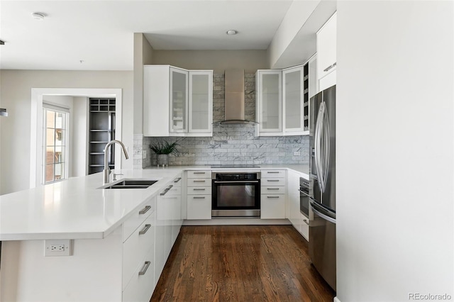 kitchen featuring a peninsula, a sink, stainless steel appliances, wall chimney range hood, and backsplash