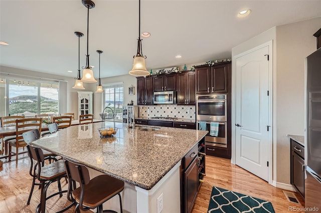 kitchen featuring light wood-type flooring, tasteful backsplash, decorative light fixtures, a center island with sink, and stainless steel appliances