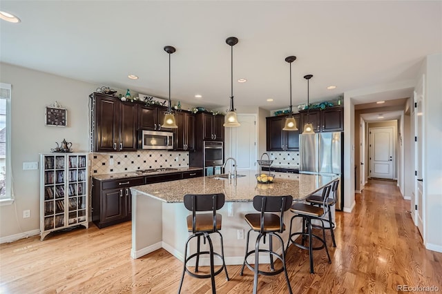 kitchen with backsplash, stainless steel appliances, light hardwood / wood-style floors, a center island with sink, and light stone countertops