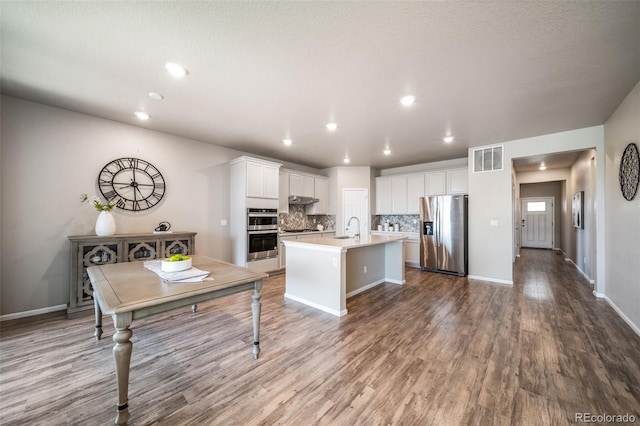 17028 Osage St, Broomfield, CO 80023 - kitchen featuring stainless steel appliances, wood finished floors, tasteful backsplash, a kitchen island with sink, and white cabinetry