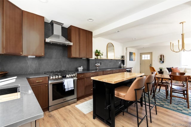 kitchen featuring wall chimney range hood, light wood finished floors, decorative backsplash, gas range, and hanging light fixtures