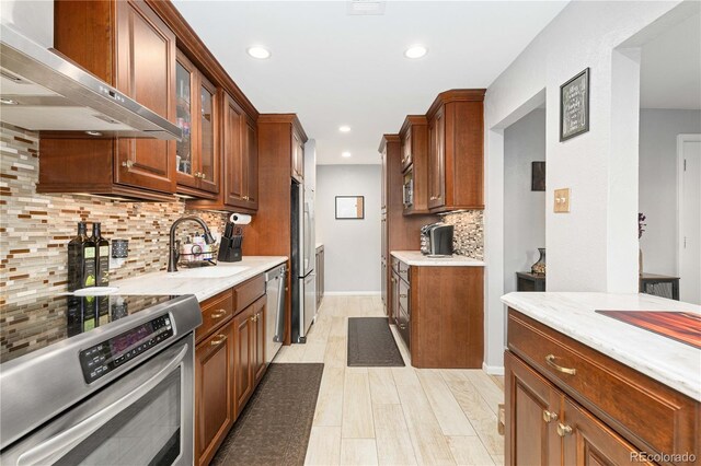 kitchen featuring a sink, appliances with stainless steel finishes, light stone countertops, wall chimney exhaust hood, and glass insert cabinets