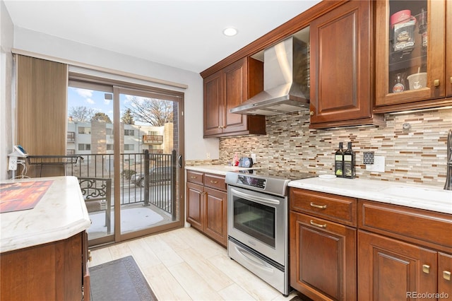 kitchen with stainless steel electric range oven, backsplash, glass insert cabinets, a sink, and wall chimney exhaust hood