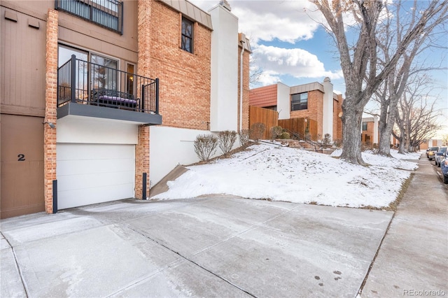 view of snow covered exterior featuring brick siding, driveway, a balcony, and an attached garage