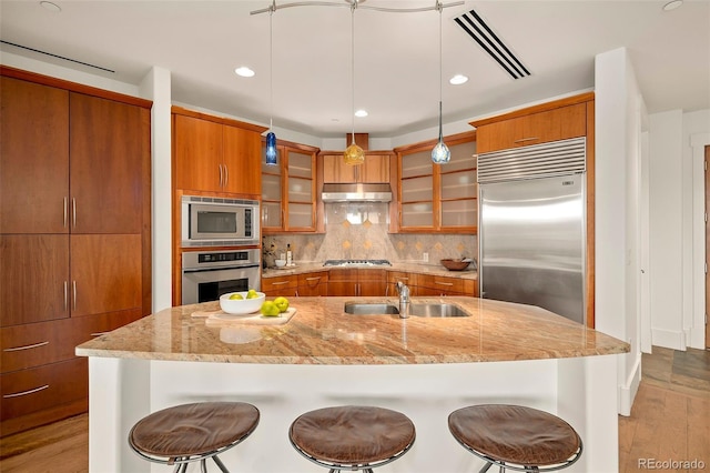 kitchen with built in appliances, brown cabinetry, a sink, and under cabinet range hood