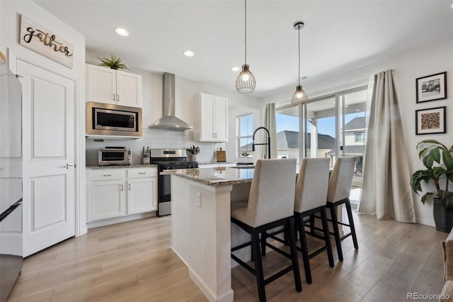 kitchen featuring a center island with sink, stainless steel appliances, white cabinets, a sink, and wall chimney range hood