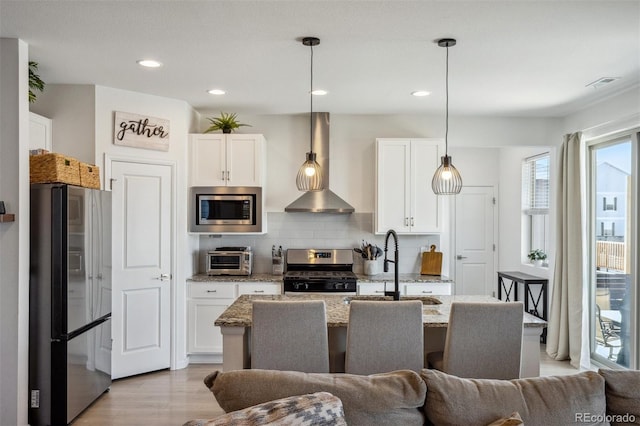 kitchen featuring light stone counters, appliances with stainless steel finishes, wall chimney range hood, and white cabinetry