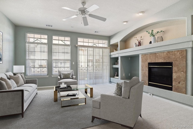 carpeted living room with ceiling fan, built in features, a tile fireplace, and a textured ceiling