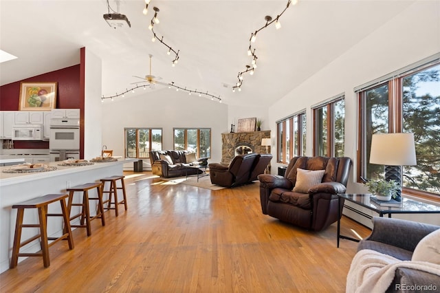 living room featuring ceiling fan, a stone fireplace, high vaulted ceiling, a baseboard heating unit, and light wood-type flooring