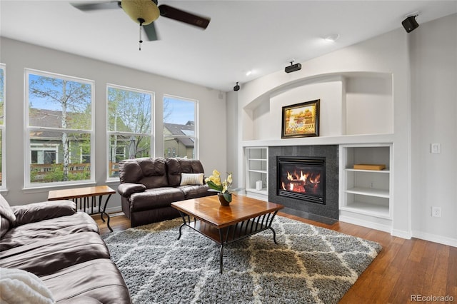 living room featuring wood finished floors, a ceiling fan, a tiled fireplace, baseboards, and built in shelves