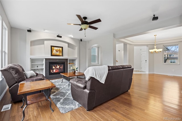 living area featuring ceiling fan with notable chandelier, visible vents, built in shelves, a tiled fireplace, and light wood-style floors
