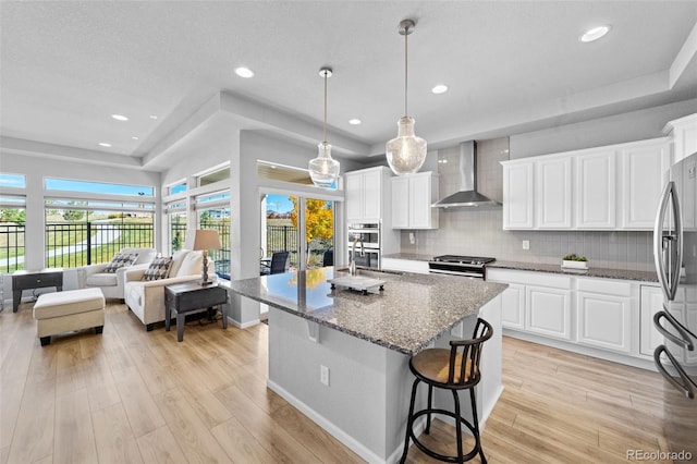 kitchen featuring wall chimney range hood, appliances with stainless steel finishes, decorative light fixtures, white cabinets, and light hardwood / wood-style flooring