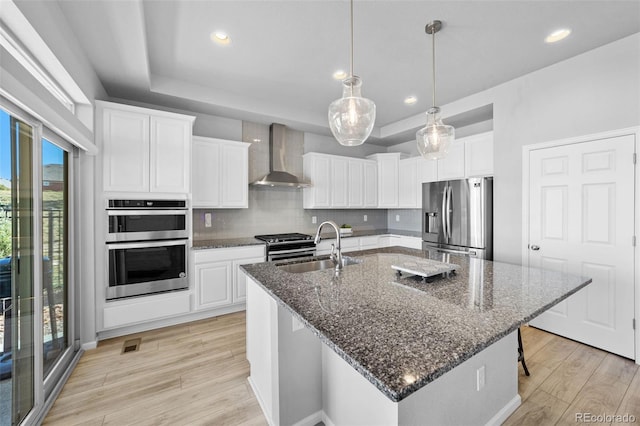 kitchen with a center island with sink, appliances with stainless steel finishes, wall chimney range hood, and light hardwood / wood-style floors