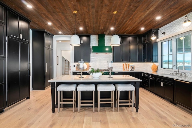 kitchen with light wood-type flooring, wall chimney exhaust hood, sink, hanging light fixtures, and an island with sink