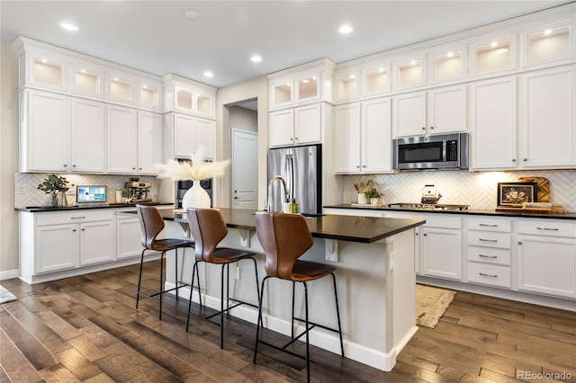 kitchen with dark countertops, dark wood-type flooring, a center island with sink, appliances with stainless steel finishes, and white cabinetry
