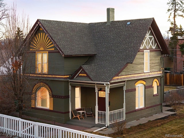 view of front of house featuring roof with shingles, brick siding, a chimney, covered porch, and fence