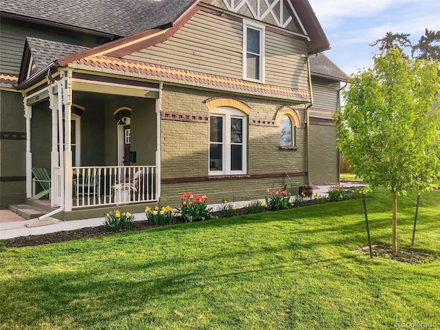 view of side of home with a yard, a shingled roof, covered porch, and brick siding