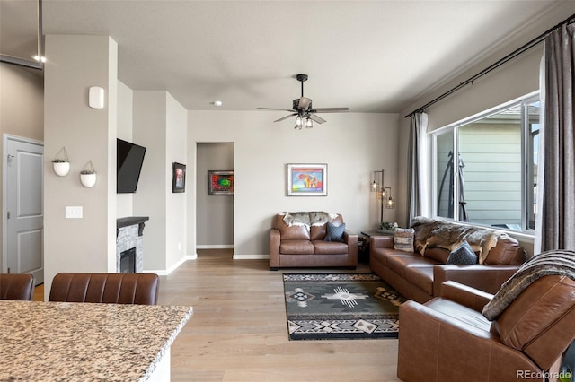 living area featuring light wood-style flooring, a ceiling fan, baseboards, and a stone fireplace