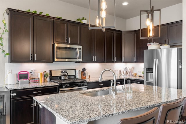 kitchen with appliances with stainless steel finishes, a sink, an island with sink, dark brown cabinetry, and light stone counters